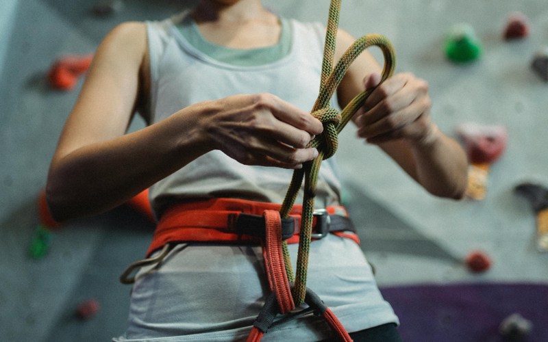 A person knots a safety harness in front of an indoor climbing wall