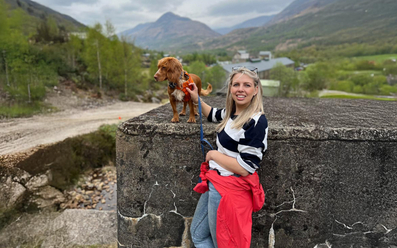 Image of Jo Bruce, a white woman with blonde hair, standing in front of a hilly landscape with the company of her dog Inka