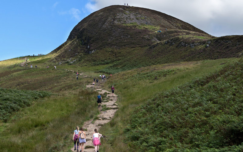 People walking up a path leading around and up a green hill under a blue sky.