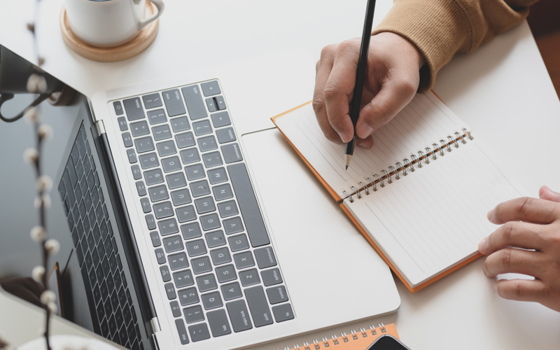 A laptop and notebook seen from above. A hand holds a pencil poised to write in the notebook.