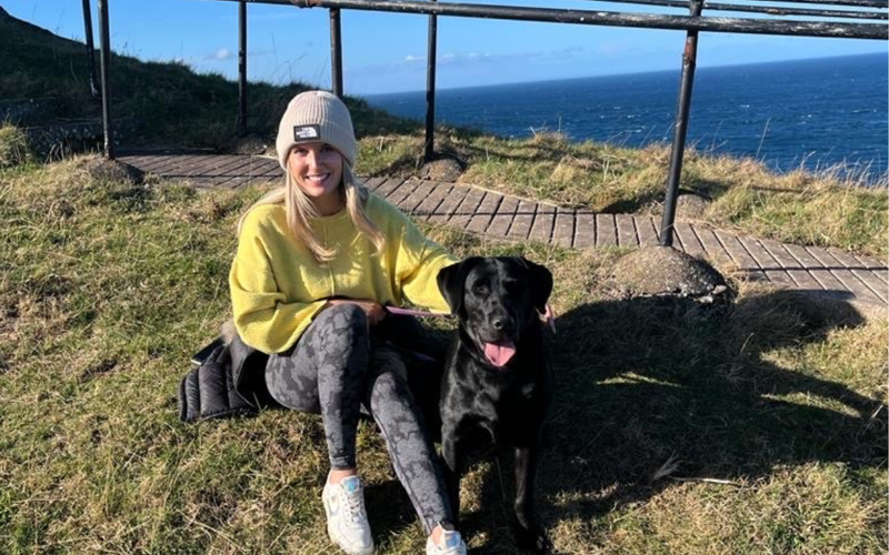 Carly McAllan poses with a black labrador, sitting on a hill with a view of the sea