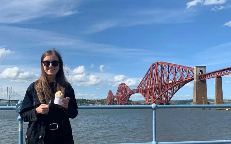 Aileen Grieve, in sunglasses and holding icecream, stands in front of a scene of the forth rail bridge